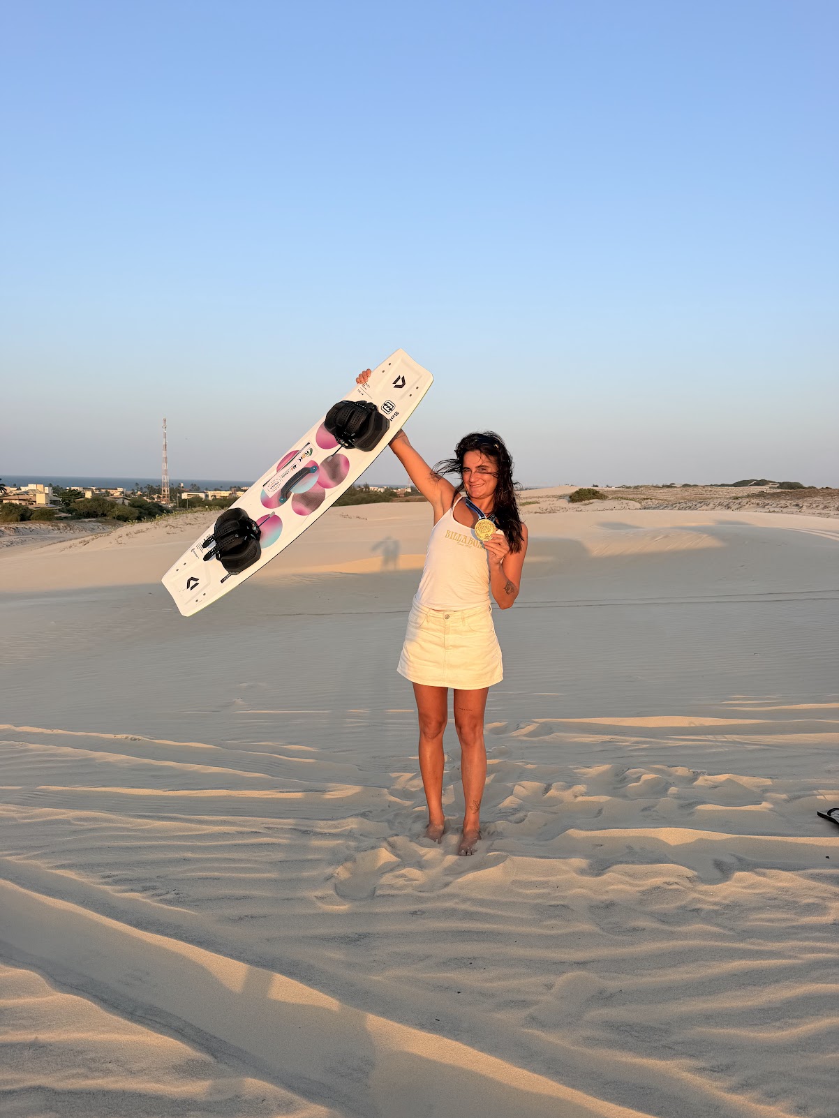 Gabi nas dunas segurando prancha e medalha de kitesurf